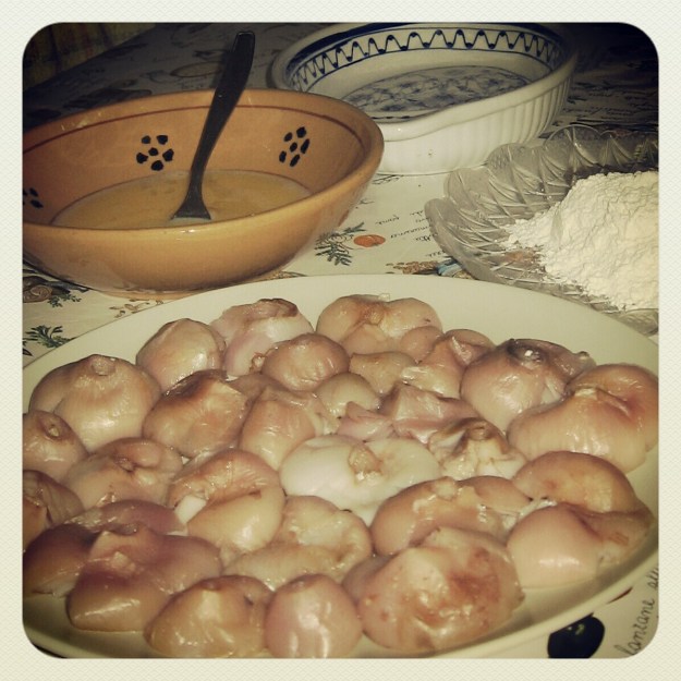 Ein Teller mit mehreren kleinen, weißen und rosa gefärbten Lampascioni, umgeben von Schalen mit geschlagenem Ei und Mehl auf einem Tisch.