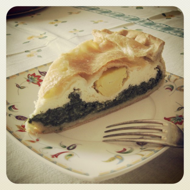 Eine Nahaufname von einem Stück Torta Pasqualina angerichtet mit einer Gabel auf einem quadratischen Teller mit florealem Dekor. Zu sehen sind deutlich die einzelnen Schichten von Teig, Spinat, Ricotta und Ei.