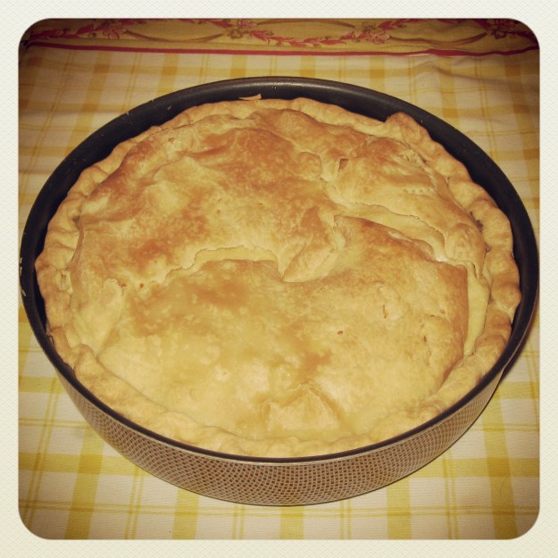 Eine goldbraune Torta Pasqualina steht in einer Metallform auf einem Tisch mit einem gelb karriertenTischtuch.