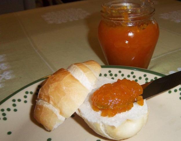 Leckere, orangefarbene Kumquatmarmelade wird von einem Messer auf ein fricshes goldgelbes Hörnchen gestrichen. Das Marmeladenglas steht neben dem Teller.