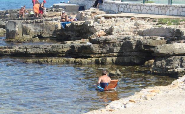 Eine Person sitzt auf einem Stuhl im Wasser in einer Badebucht zwischen Felsen.