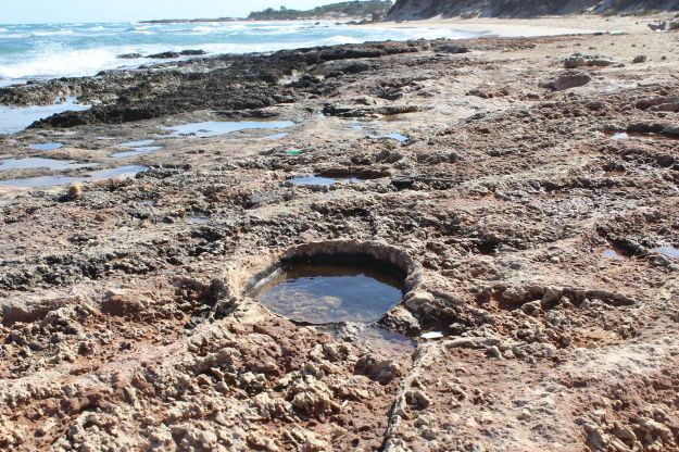 Ein felsiger Strand des Naturschutzgebietes von Torre Guaceto in Apulien zeigt ein kreisrundes, geheimnisvolles Loch, dass mit glasklarem Meerwasser gefüllt ist.