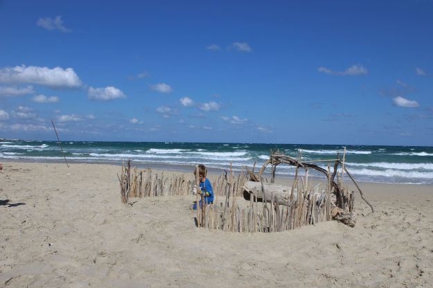 Ein Kind spielt in einer improvisierten Festung aus Treibgut am hellen Sandstrand des Naturschutzgebietes von Torre Guaceto in Apulien, im Hintergrund rollt das grünblaue Meer in schaumbesetzten Wellen an den Strand.