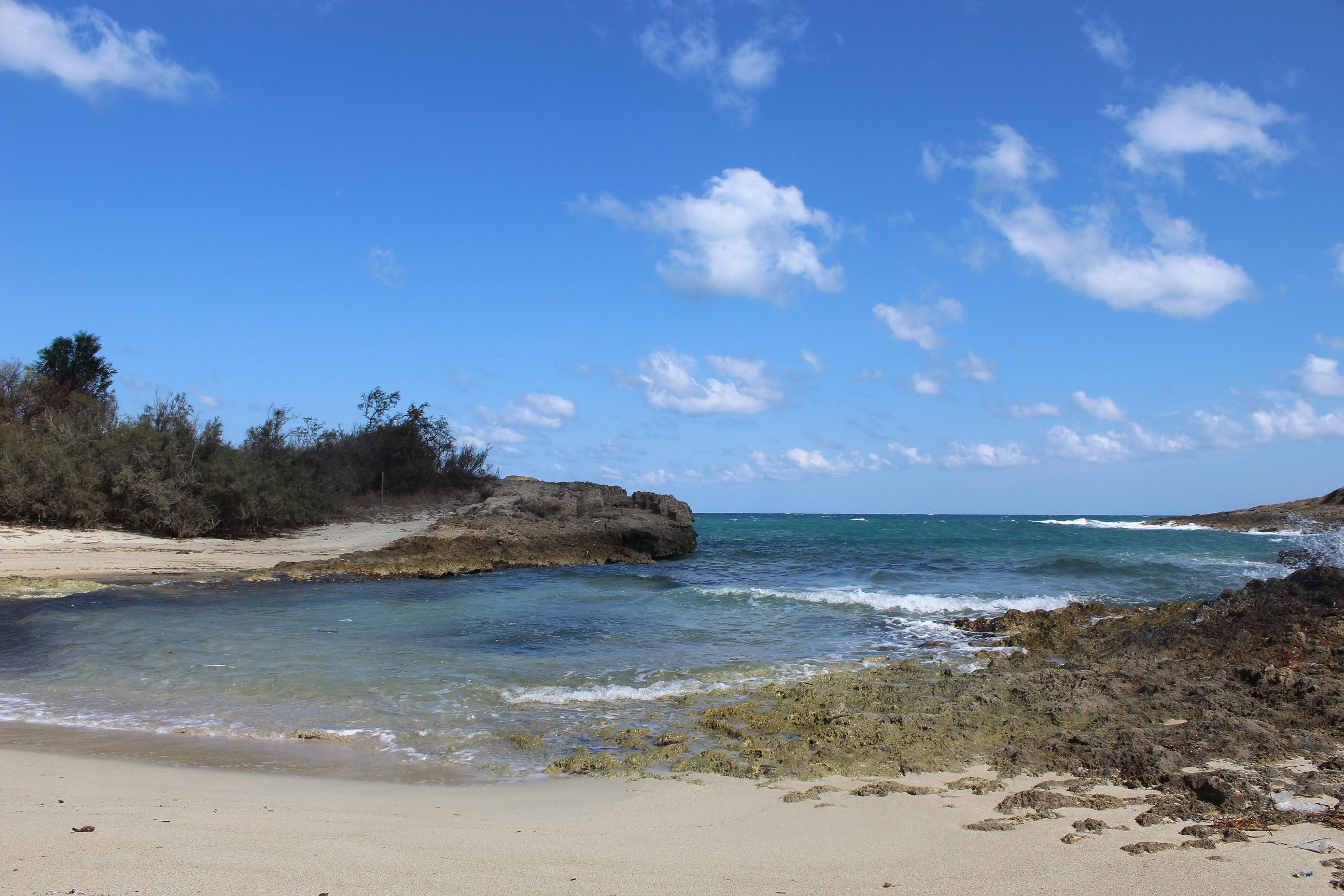 Eine Meeresbucht mit hellem Sandstrand wird eingeschlossen von felsigen Abschnitten undklares blaues Wasser rollt in niedrigen Wellen an den Strand des Naturschutzgebietes von Torre Guaceto in Apulien.