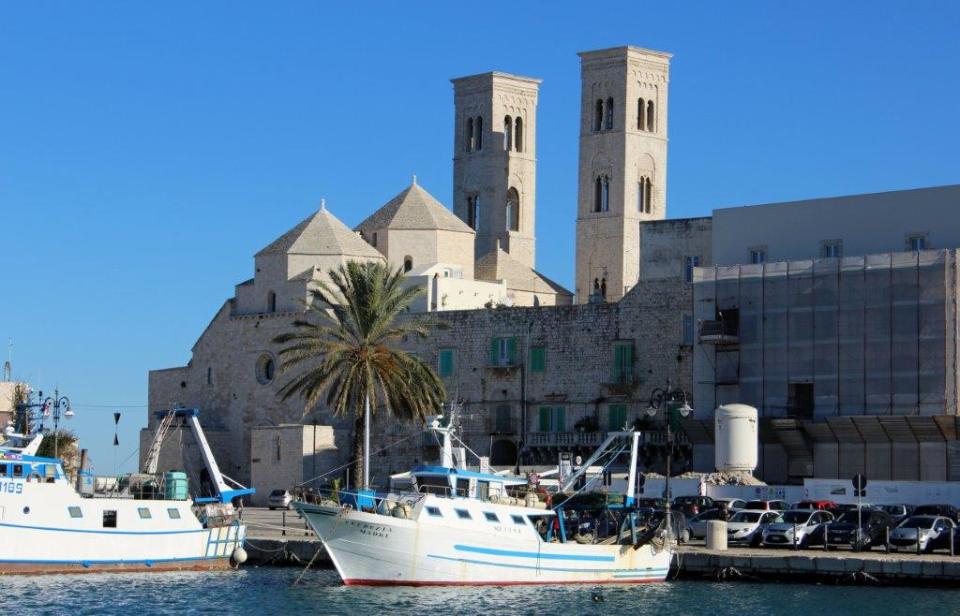 Im Hafen von Molfetta, Apulien liegen zwei Fischerboote, hinter denen sich die Kathedrale aus hellem Sandstein erhebt.