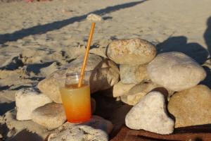 Ein orange-gelber Cocktail mit Strohhalm steht auf glatt gewaschenen Steinen an einem Sandstrand.