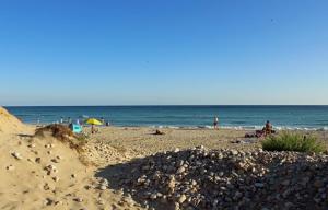 Eine sandige Düne mit kleinen Steinchen gibt den Blick frei auf wenige Badegäste am Sandstrand der Malediven des Salento. Dahinter das blaue Meer.