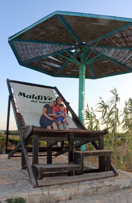 Eine dreiköpfige Familie posiert auf dem überdimensionalen Liegenstuhl, Wahrzeichen des Strandes "Malediven des Salento" unter einem genauso überdimensionalem Sonnenschirm.