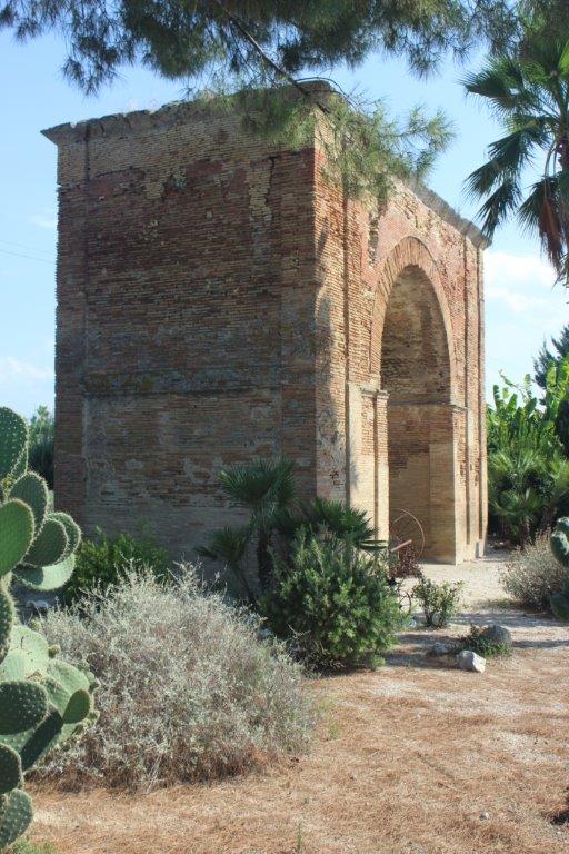 Inmitten der Ausstellungsfläche eines Gartenbaubetriebs erhebt sich der Torbogen Archo Varrone Archo Varrese auch Archo Traiano genannt, in Canosa zwischen kleinen Büschen und Kakteen.