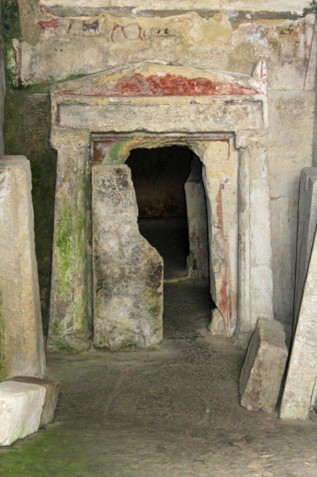 Ein hohes Tor mir Steintüren und rudimentär erkennbaren Fresken bildet den Eingang zur Grabanlage Ipogeo Scocchera B-1 in Canosa, Apulien.