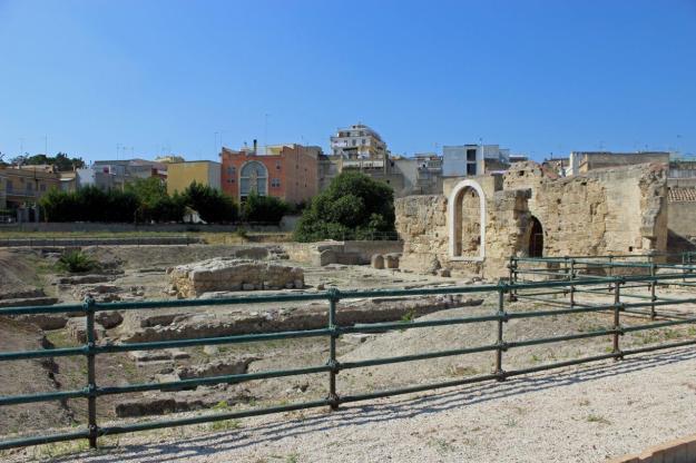 Vor den modernen Wohnhäusern der süditalienischen Stadt Canosa, liegt die mit einem niedrigen Zaun umgebene Ausgrabungsstätte „Parco Archeologico di San Giovanni“. Rechts erheben sich andeutungsweise die Mauern der Täuferkirche des Heiligen Johannes.