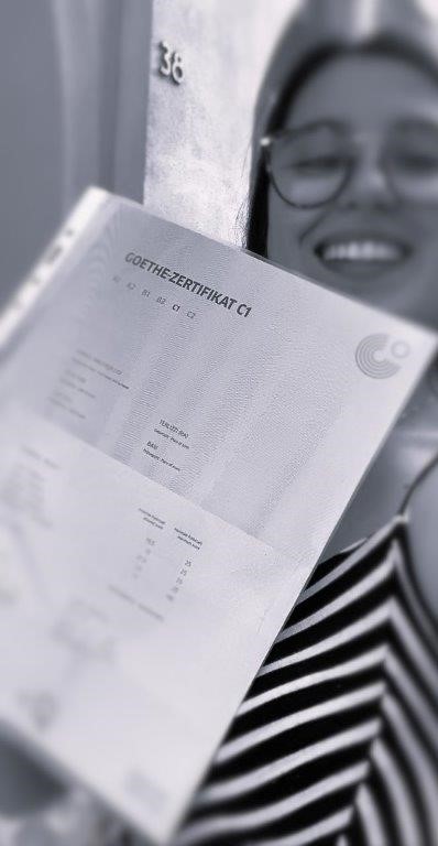 Eins stolze Deutschschülerin zeigt lächelnd ihr C1 Goethe Zertifikat in die Kamera, das sie nach einem Vprbereitungskurs mit Corinna bestanden hat.