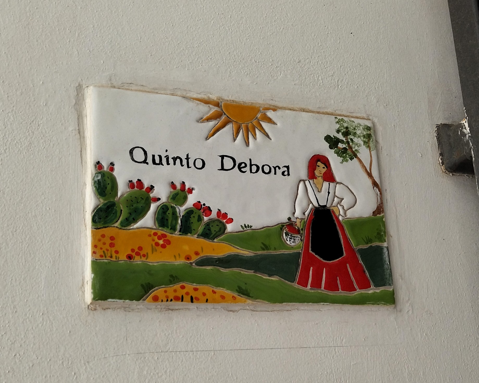 Türschild aus Keramik zeigt eine Frau im traditionellen Kleid Pacchiana auf einem Feld. Im Hintergrund sieht man Kaktusfeigen. Das Schild stammt von Anna Maria Pagliei einer Künstlerin aus Pisticci in Basilikata, Süditalien