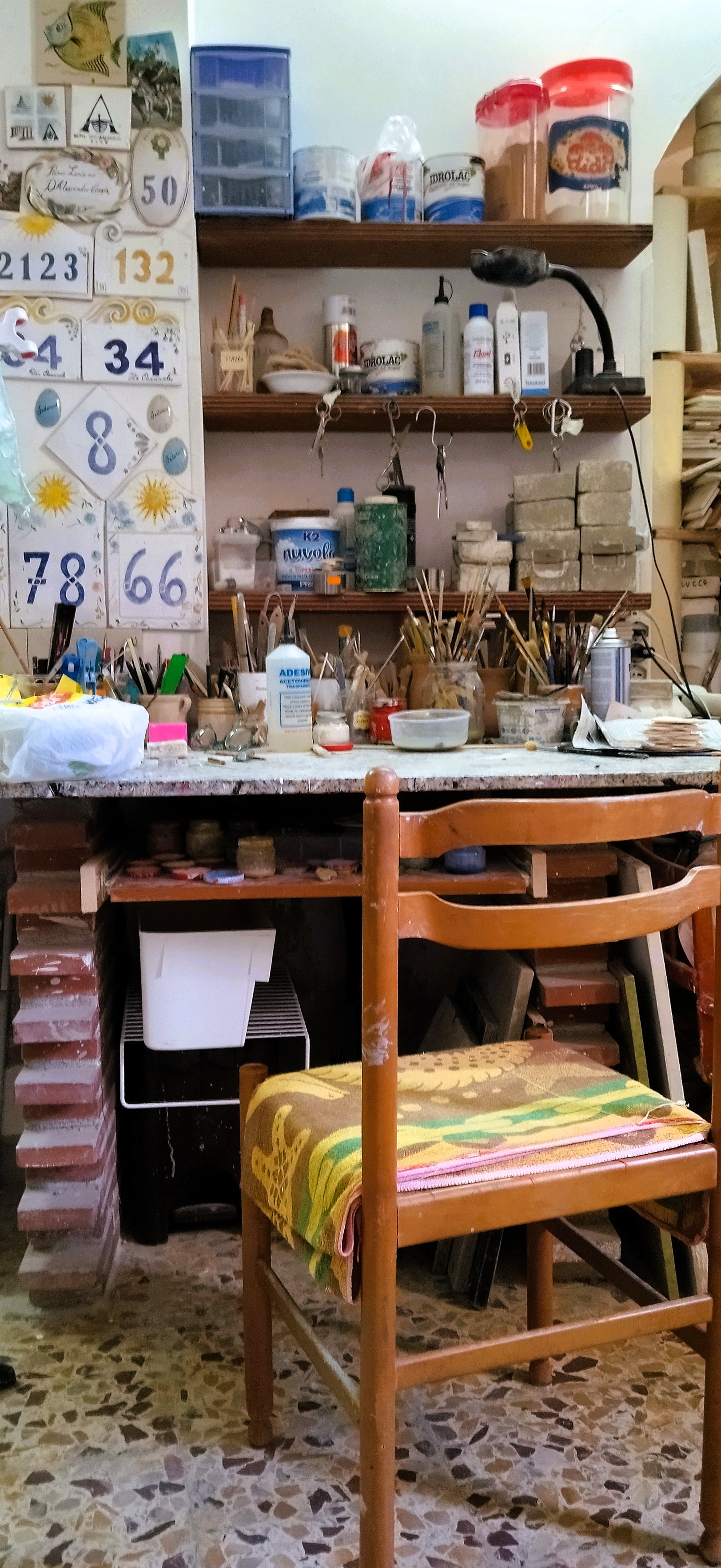 Ein Holzstuhl steht vor einem Arbeitstisch mit Regalen und vielen Gefäßen, Pinseln und Schildern in der Keramikwerkstatt der Künstlerin Annamaria Pagliei in Pisticci Basilikata.