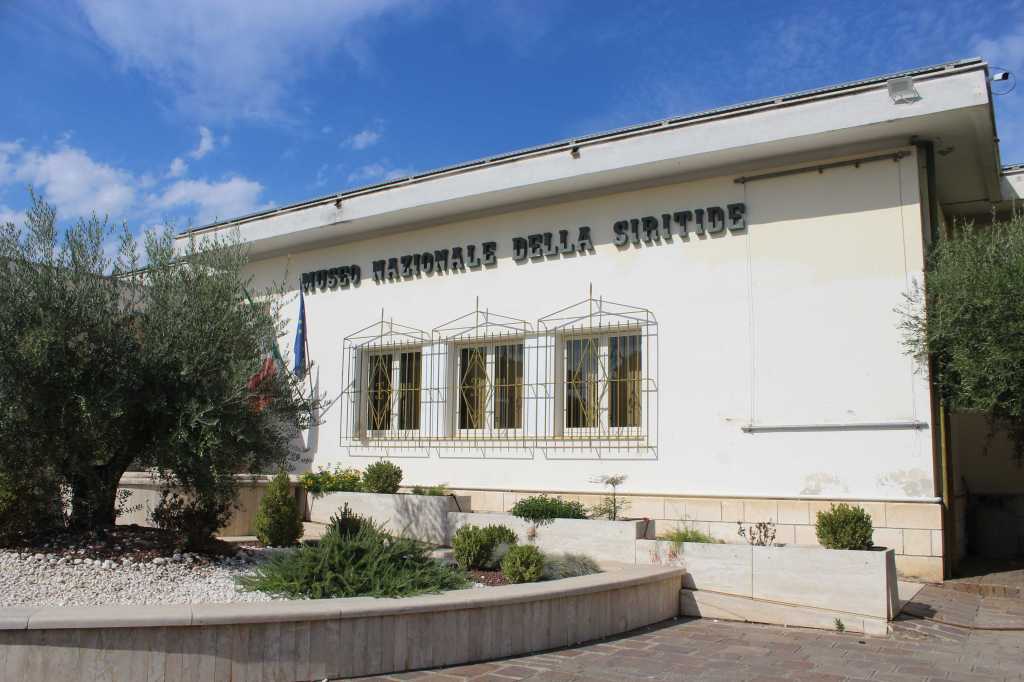 Der Eingang des Archäologische Nationalmuseums Siritide in Policoro, Basilikata, liegt hinter einer Rabatte mit niedrigen Sträuchern und einem kurz gehaltenen Olivenbaum.