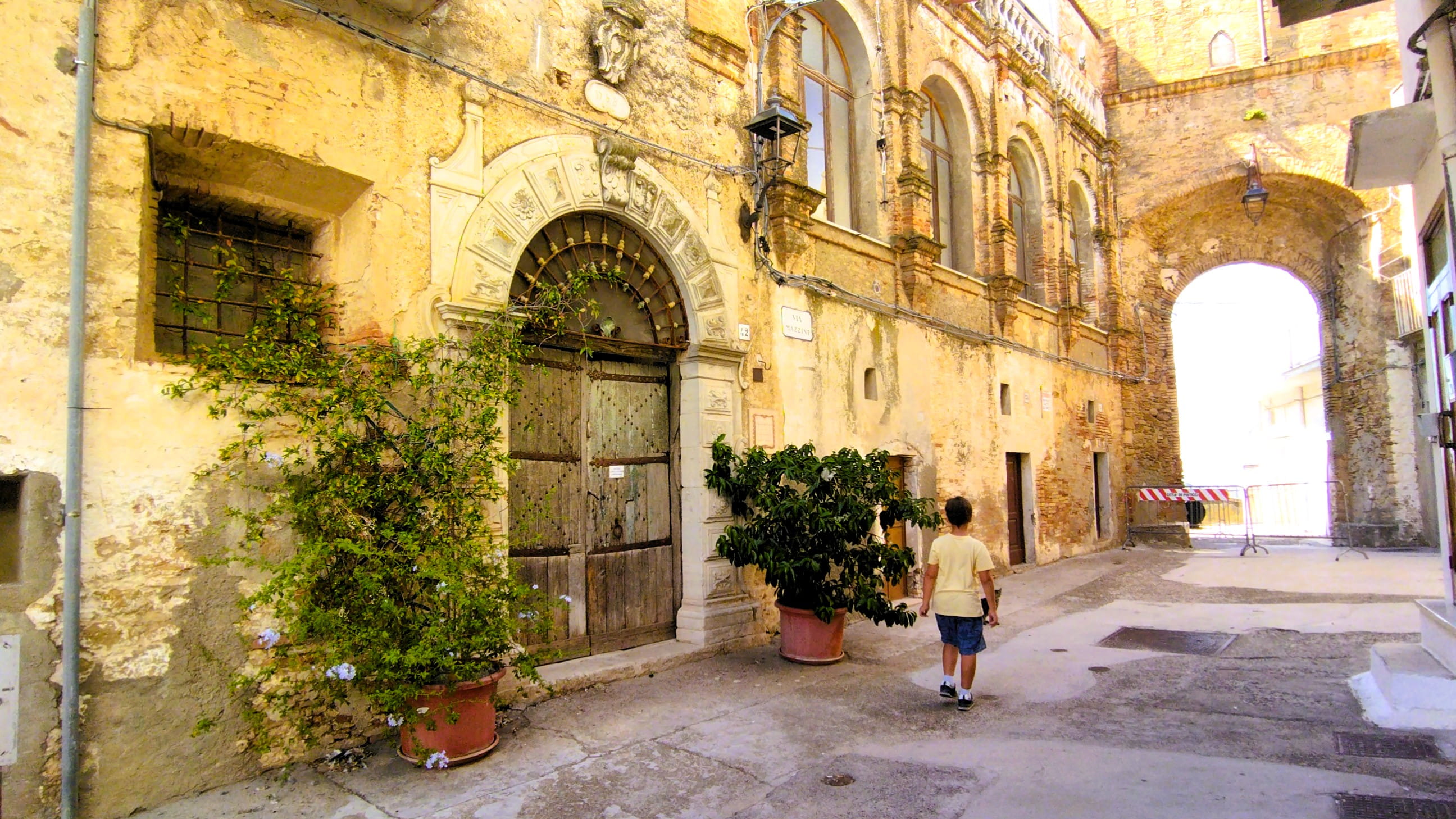Das Bild zeigt einen in die Jahre gekommenen historischen Palazzo in Pisticci, der vermutlich Teil des Kastells war und vor dem eine Straße zu einem vom Sonnenlicht hell erleuchtetn Durchgang führt.