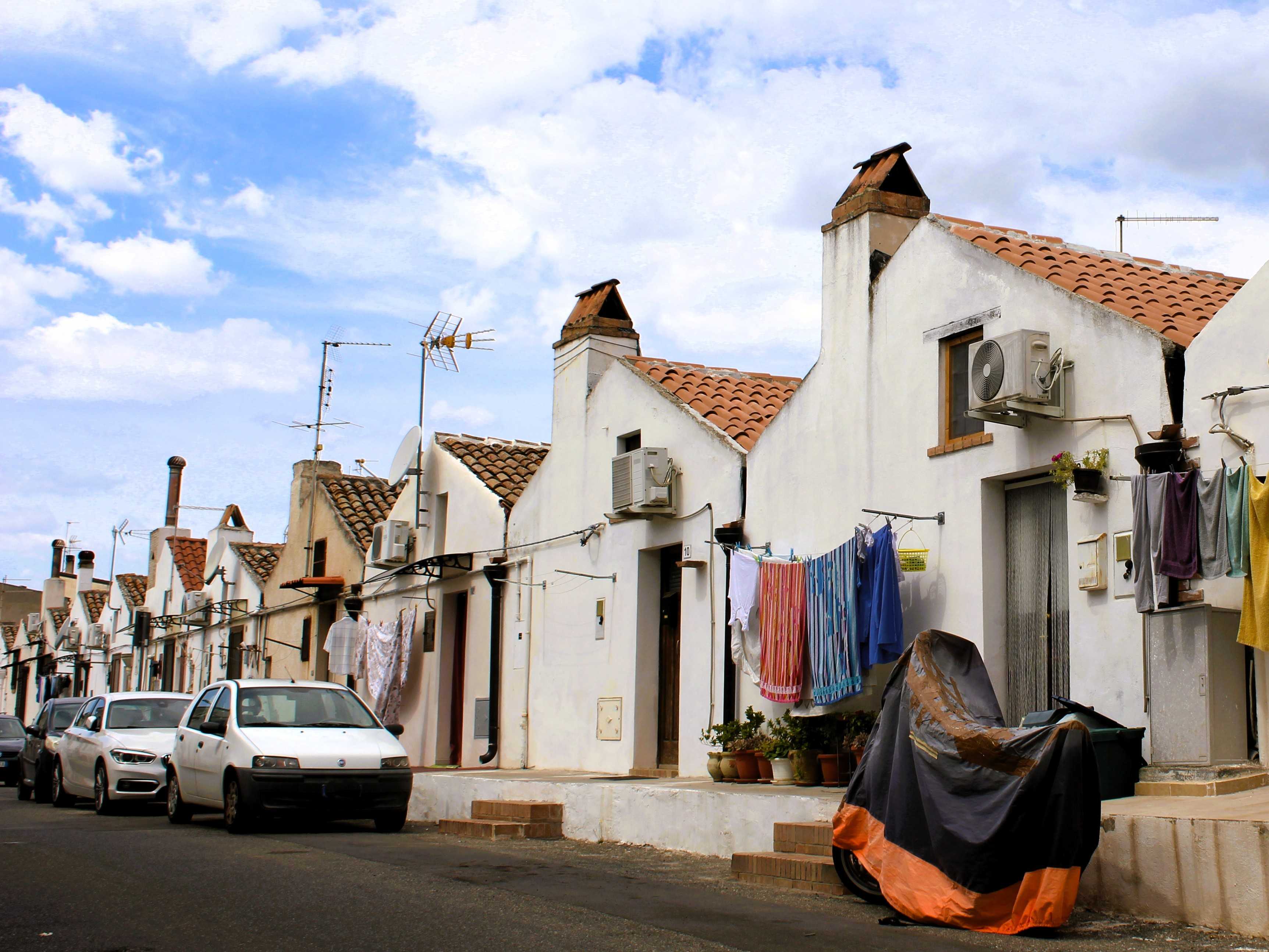 Eine für Basilikata ungewöhnliche Reihenhausstraße mit Häusern mit Spitzdach in Policoro. Davor hängt Wäsche auf Wächeleinen und am Straßenrand parken Autos sowie ein Motorrrad.