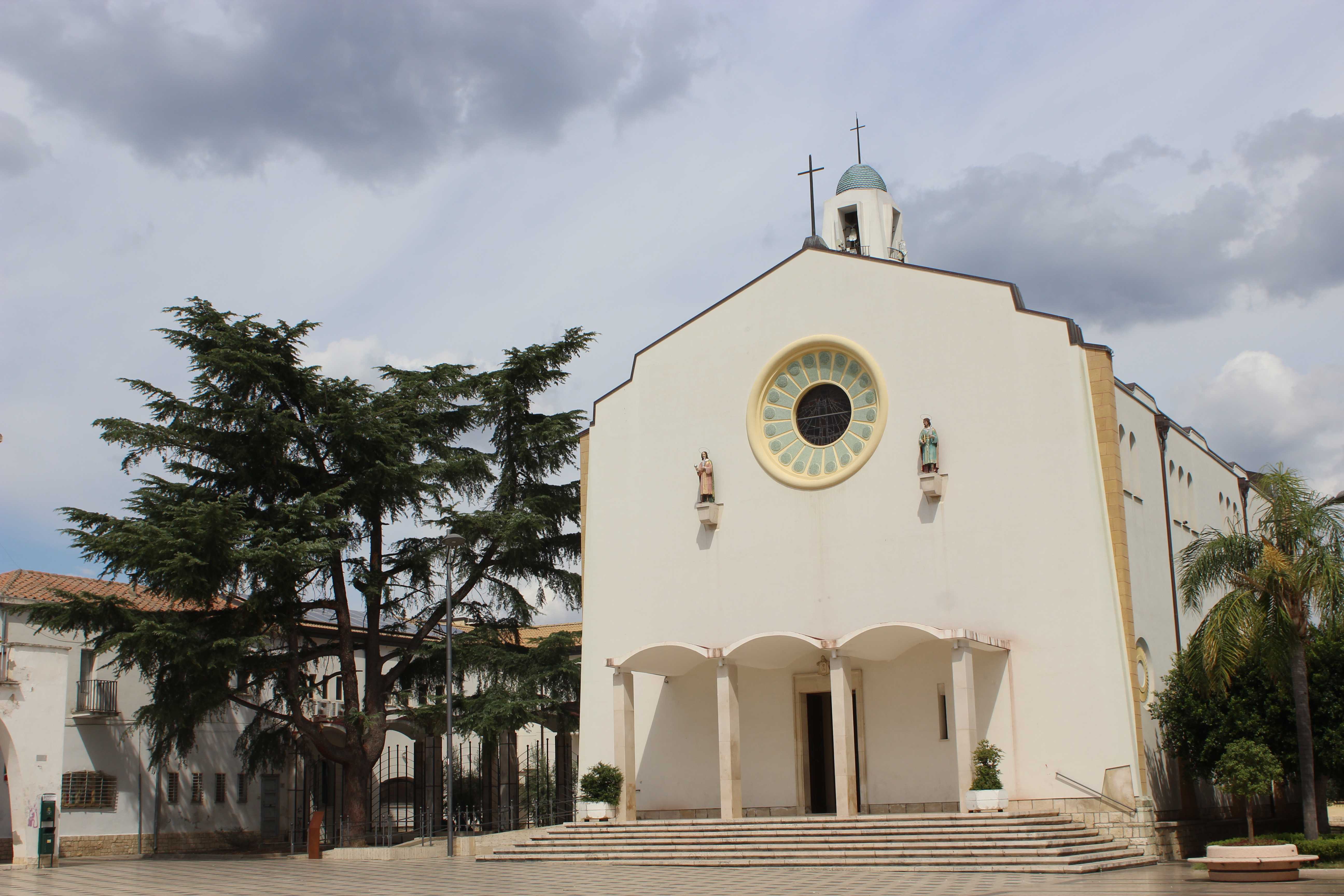 Die weiße Kirche der Santa Maria d'Anglona erhebt sich auf einem Platz flankiert von Nadelbäumen und Palmen in Policoro.