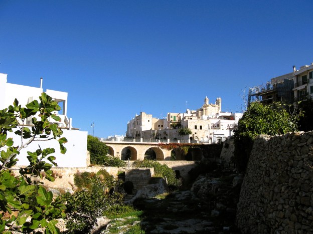 Hinter der grün bewacshenen Lama, die zum Stadtstrand von Polignano a Mare in Apulien führt erhebt sich hinter der mittelalterlichen Brücke die Altstadt.