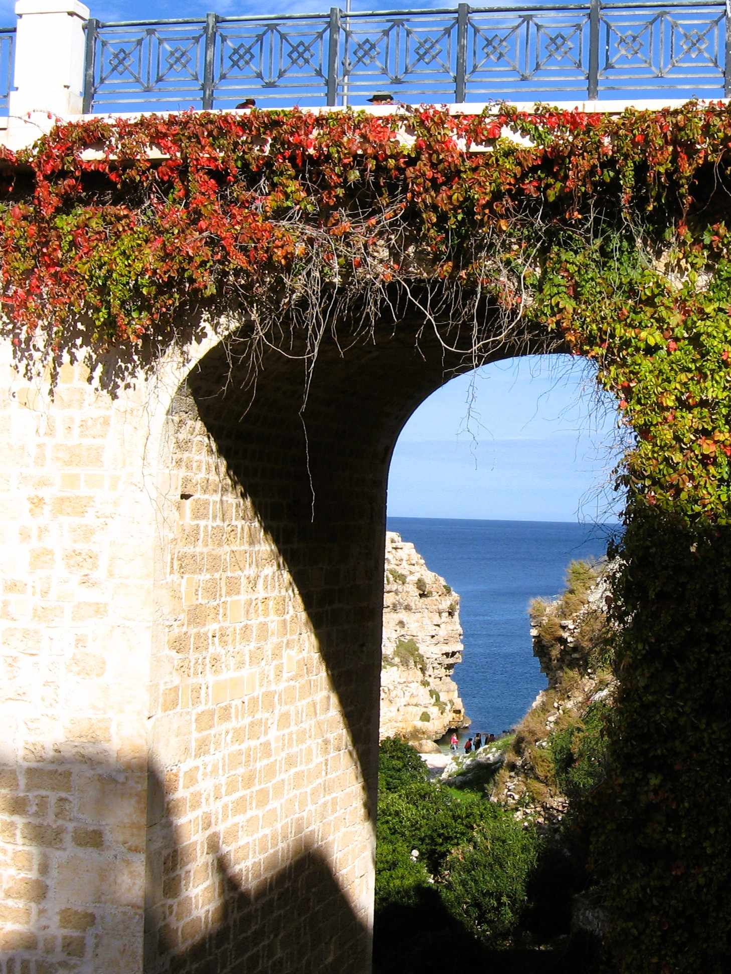 Zwischen zwei Pfeilern einer bewachsenen Brücke hindurch sieht man den Stadtstrand von Polignano a Mare und das Meer zwischen zwei Klippen liegen.