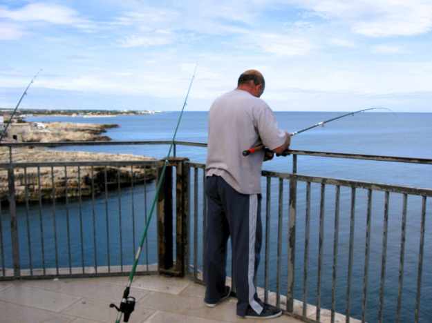 Ein Angler kontrolliert seine Angel auf einem Aussichtsbalkon in Polignano a Mare, während zwei andere Angeln auf das blaue Meer hinaus zeigen.