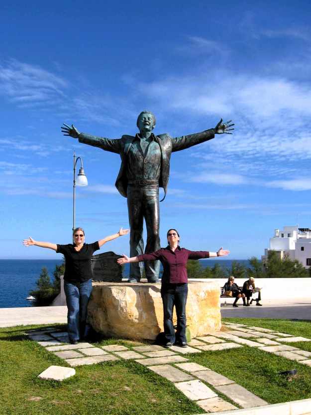 Auf der der Altstadt gegenüber liegenden Seite Polignanos steht das Denkmal für den berühmten italienischen Sänger Domencio Modugno der direkt am Meer seine Arme ausbreitet. Davor stehen zwei Touristinnen in der gleichen Haltung.