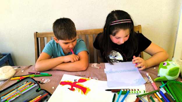 Ein Junge und ein Mädchen sitzen auf einer Holzbank am Tisch und malen. Auf dem Tisch liegen viele Stifte, zwei Federtaschen und das Malpapier.