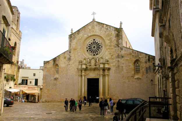 Das Foto zeigt die Sandsteinkathedrale von Otranto mit ihrem offenen Platz und einigen Touristen.