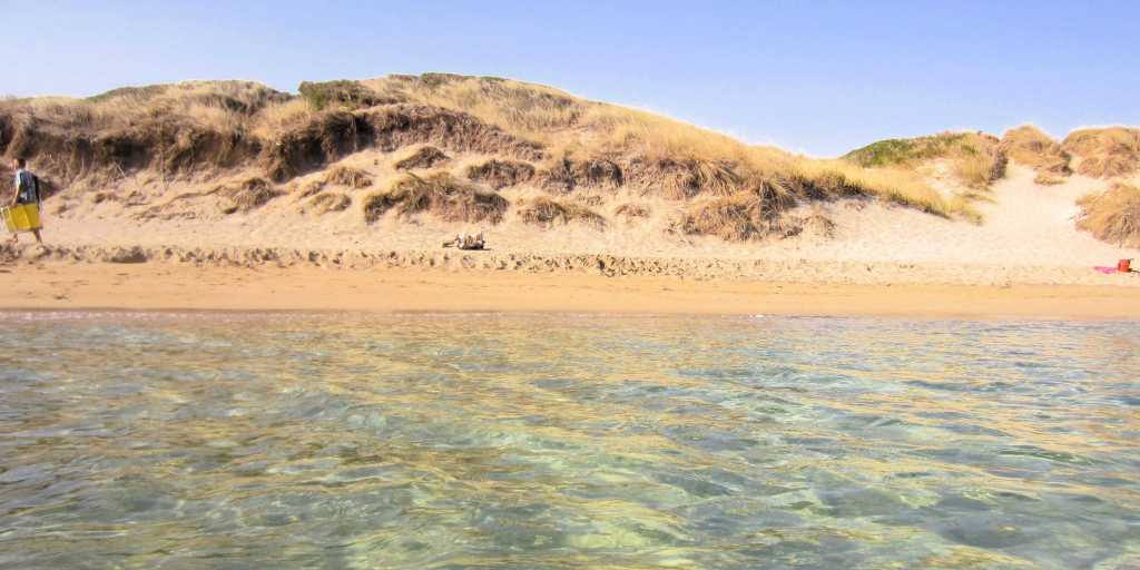 Glasklares Meerwasser am Strand von Ostuni, hinter dem sich eine bewachsene Düne erhebt