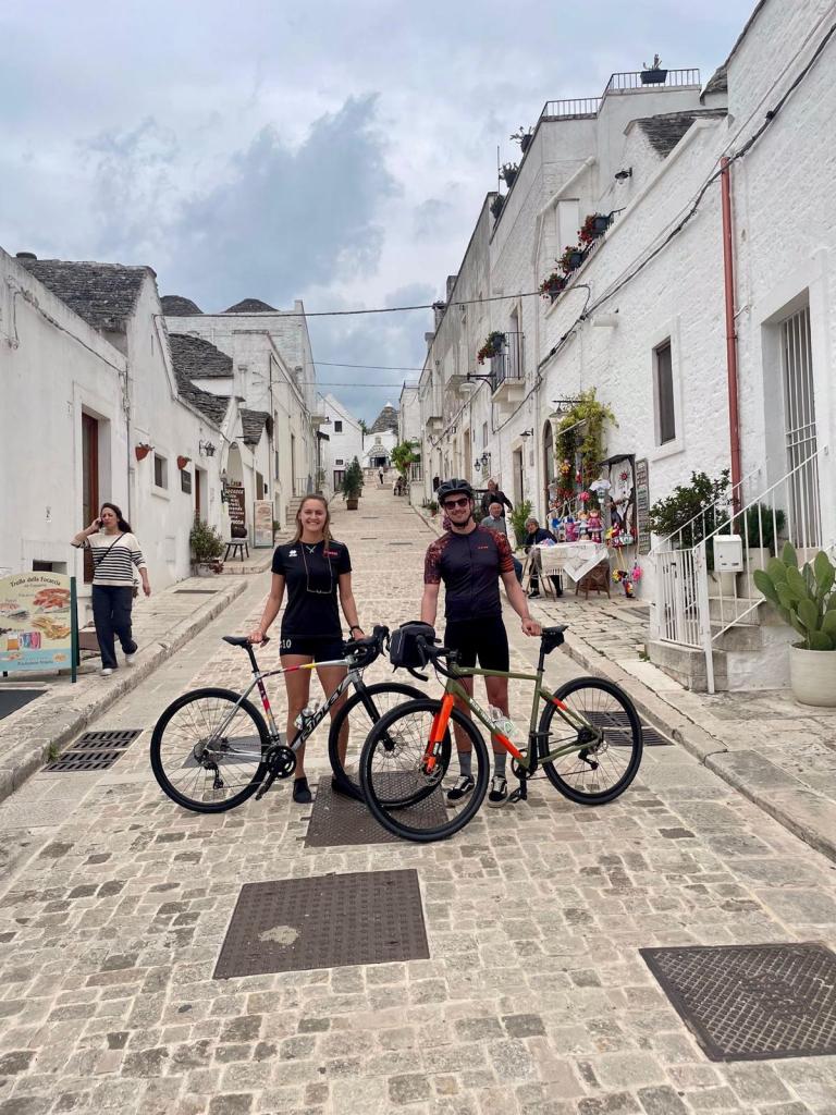 zwei Radfahrer auf einer gepflasterten Straße in einer weißen Stadt im Valle d'Itria in Apulien.