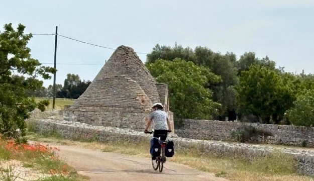 Ein Radfahrer fährt auf einer schmalen Straße im Hinterland Apuliens auf eine Kurve mit einem verfallenden Trullo zu.