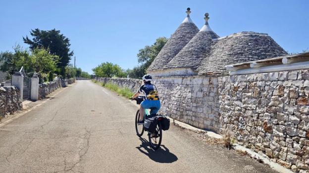 Ein sportlicher Radfahrer mit Satteltaschen fährt über eine schmale Straße zwischen Trockenmauern an Trullis im Hinterland von Apulien vorbei.
