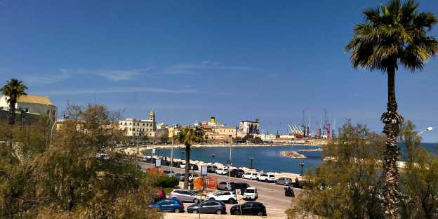 Blick von der Stadtmauer in Bari auf den Altstadtbogen, im Vordergrund eine Palme und weitere Bäume. Im Hintergrund die Basilika und hinter der Altstadt ein Kreuzfahrtschiff.