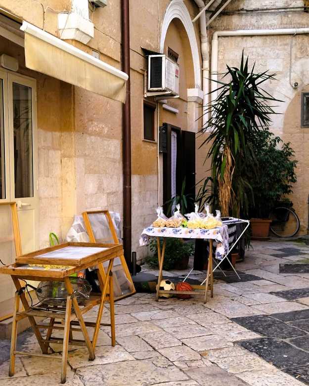Siebe zum Trocknen von frischer Pasta vor einer Hauswand in der Altstadt von Bari, daneben abgepackte Pasta auf einem kleinen Verkaufstisch.