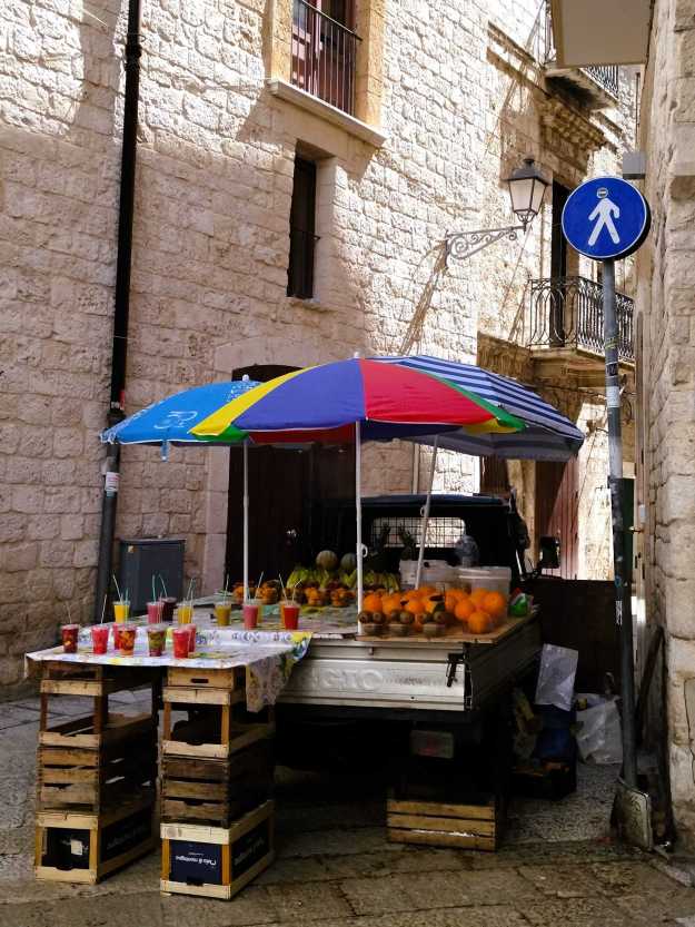 Ladefläche eines Kleintransporters mit Orangen, Bananen und bunten Saftbechern als improvisierter Saftstand in der Altstadt von Bari, überdacht von drei bunten Sonnenschirmen.